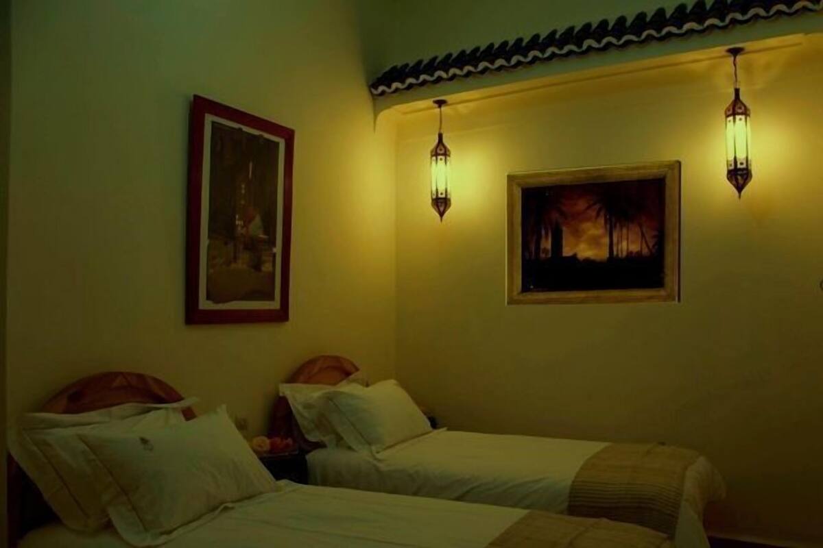 A room with two beds, a painting on the wall, and two wall-mounted lamps.