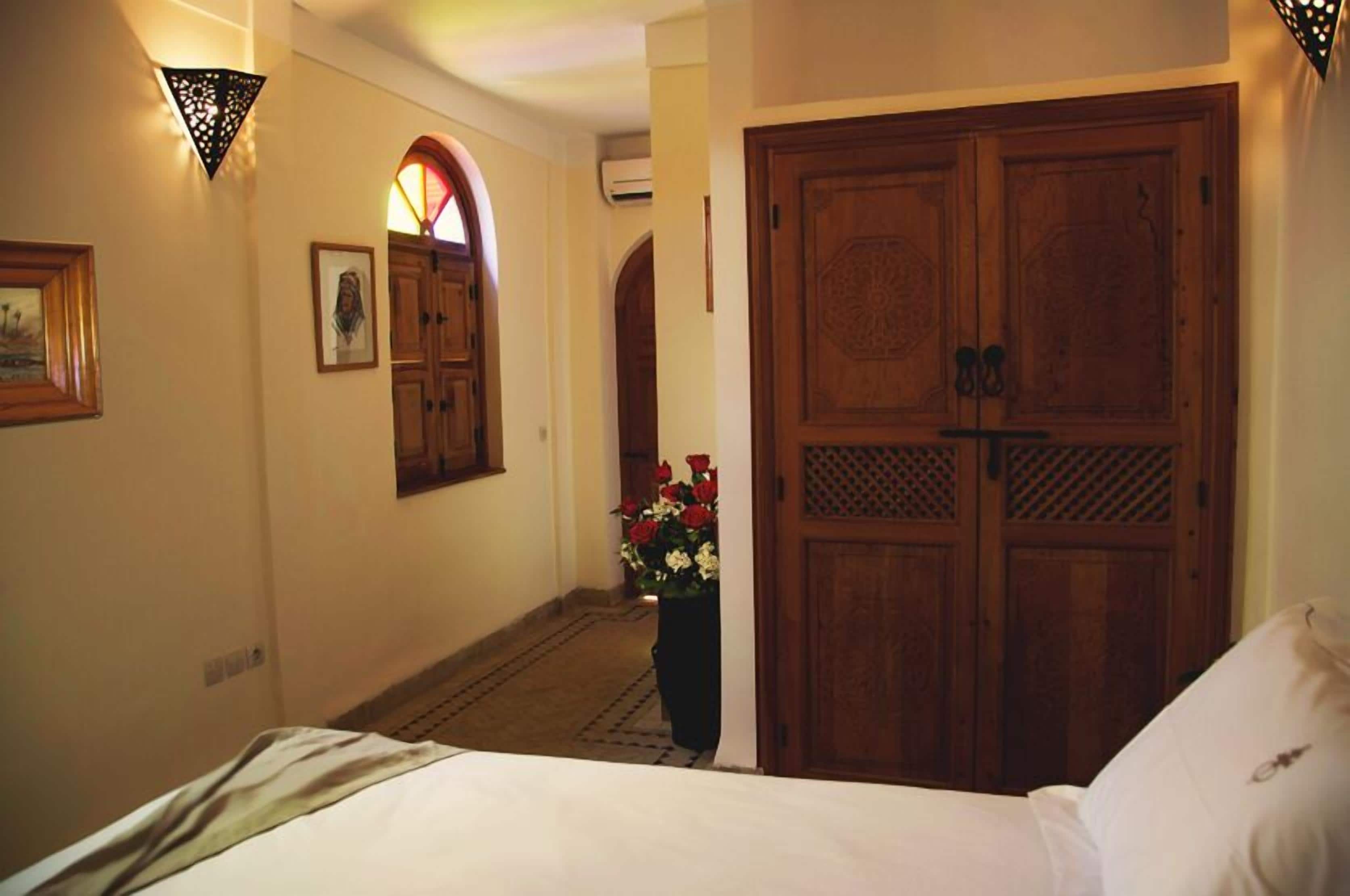 A hotel room with a bed, a wooden wardrobe, a wall-mounted light fixture, and a framed picture.