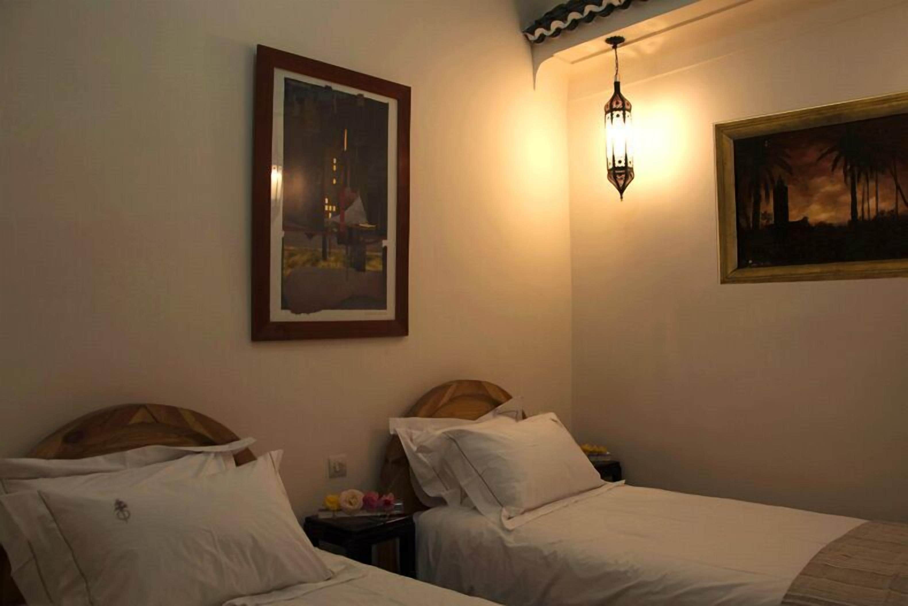 A hotel room with two beds, a nightstand, a wall-mounted lamp, and two framed pictures.