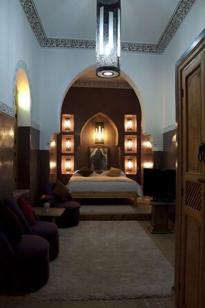 A bedroom with a large bed, a TV, and a seating area with ottomans. - Riad Granvilier (Marrakech)