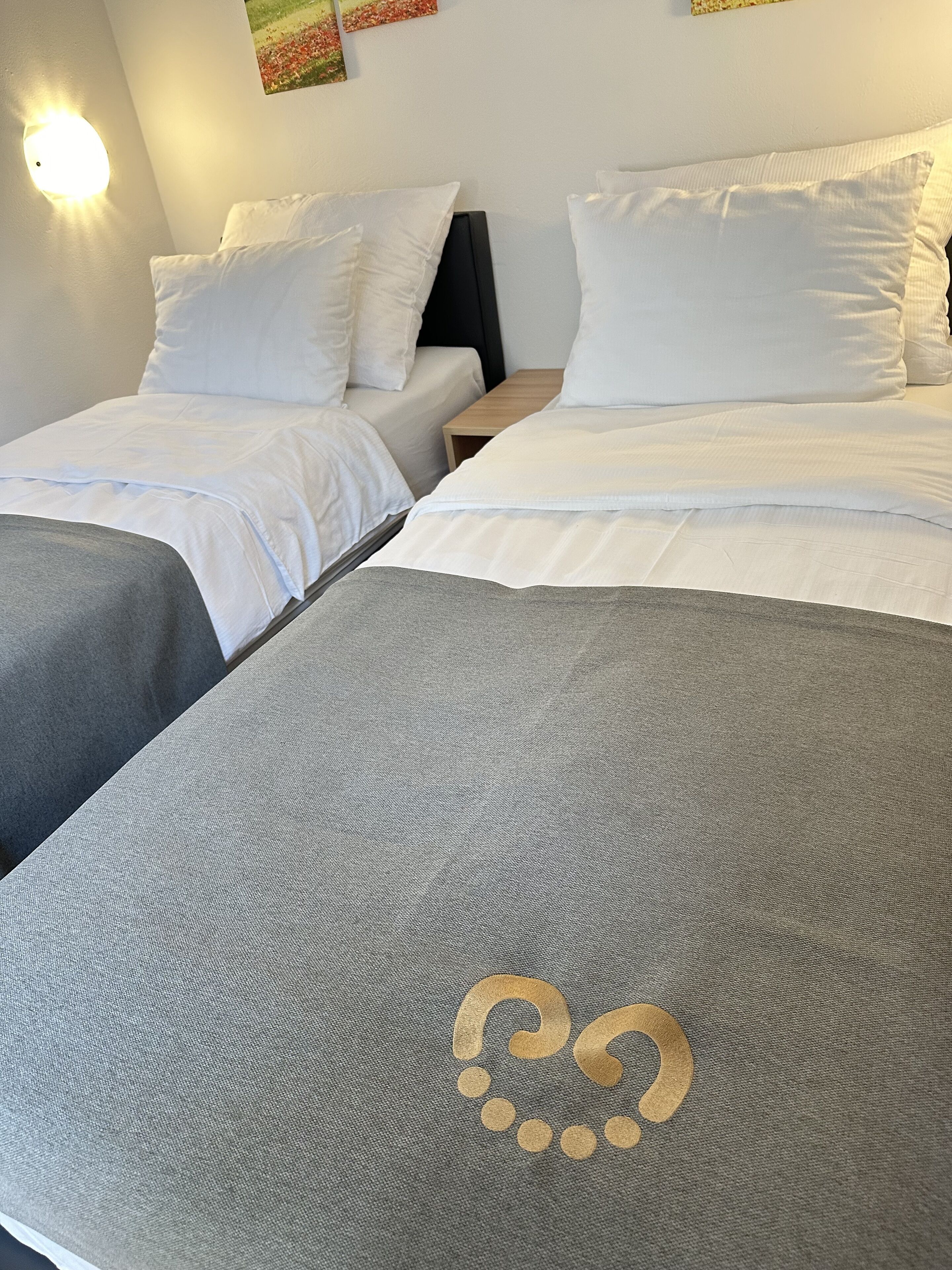 A hotel room with two beds, each with white bedding and pillows. There is a small wooden nightstand between the beds.
