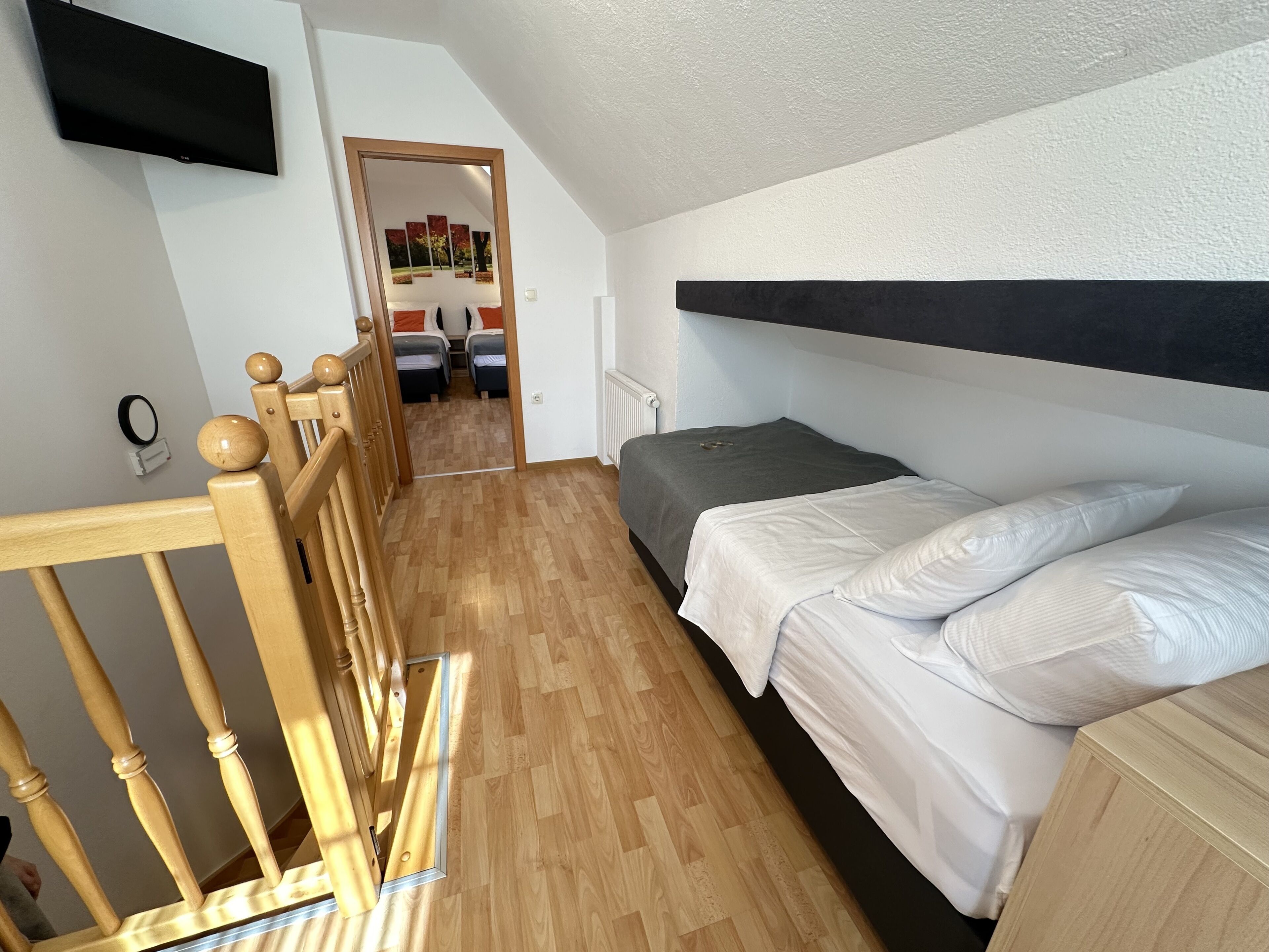 Een slaapkamer met een houten bedframe, een grijze matras en witte beddengoed. Er is een houten hoofdbord, een aan de muur bevestigde televisie en een deur naar een andere kamer.