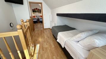 Een slaapkamer met een houten bedframe, een grijze matras en witte beddengoed. Er is een houten hoofdbord, een aan de muur bevestigde televisie en een deur naar een andere kamer.
