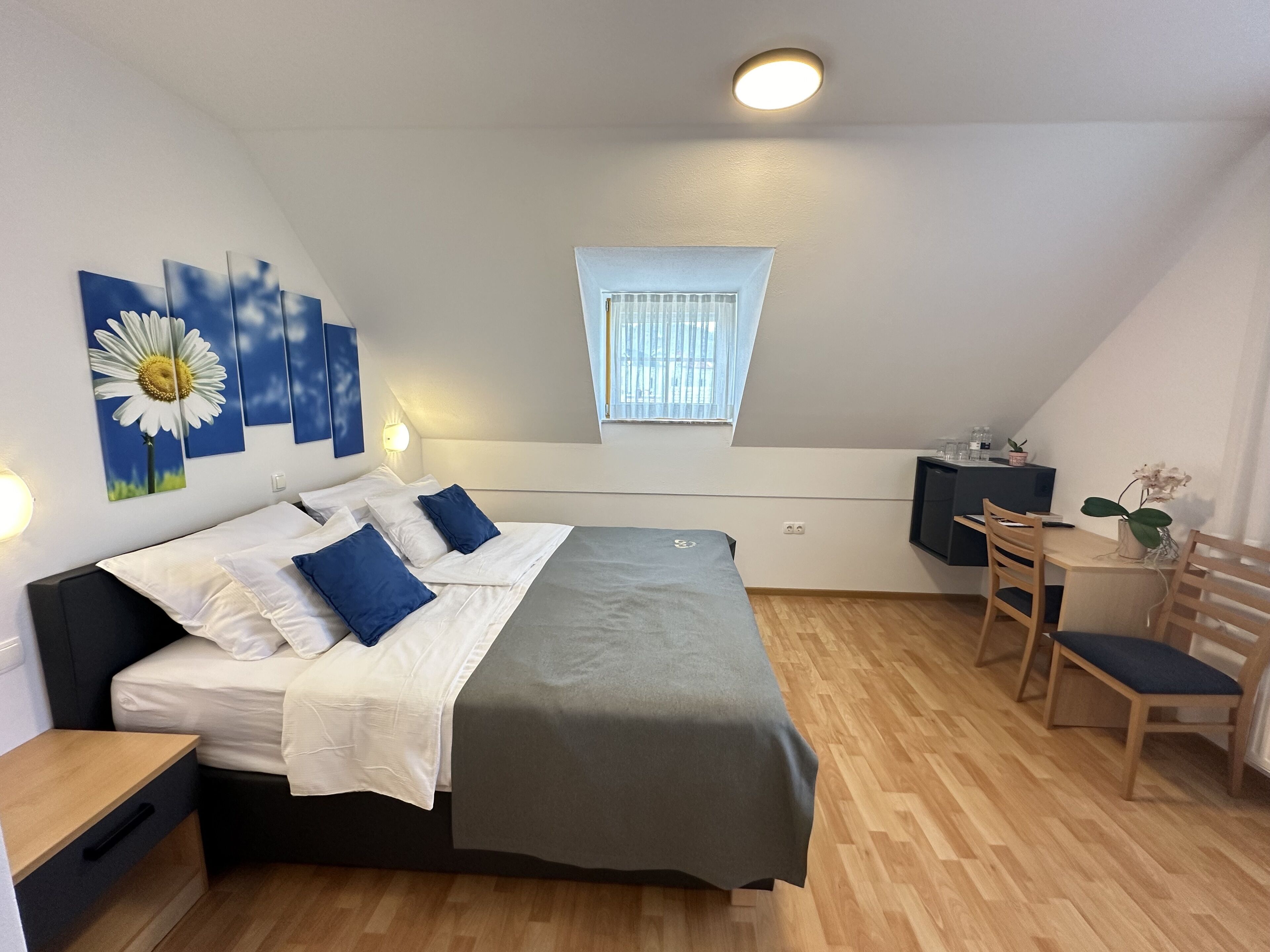 A bedroom with a bed, bedside tables, a desk, a chair, a window, and wall art.