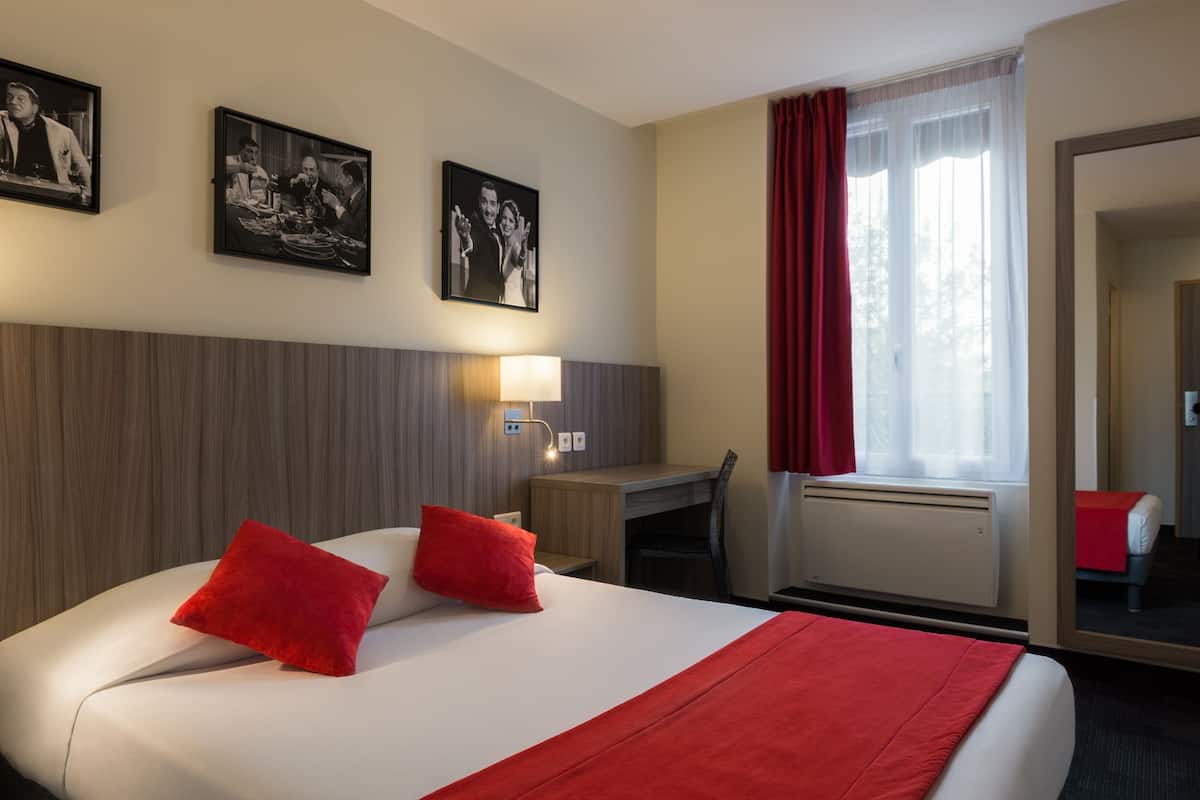 A hotel room with a bed, a desk, a chair, a lamp, and two framed pictures on the wall.