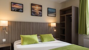 A hotel room with a bed, two green pillows, a nightstand, a wall-mounted lamp, and a wardrobe. - Reims Hotel (Paris)