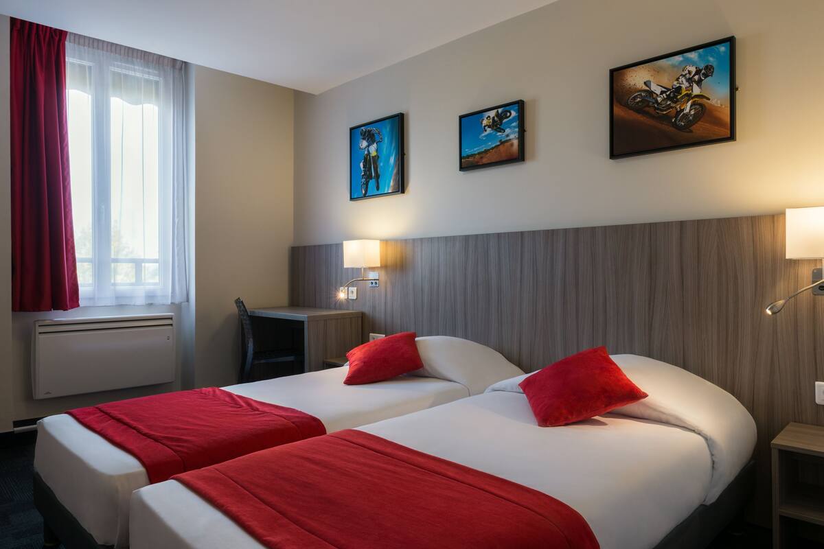 A hotel room with two beds, red bedding, a desk, a chair, and wall-mounted artwork.