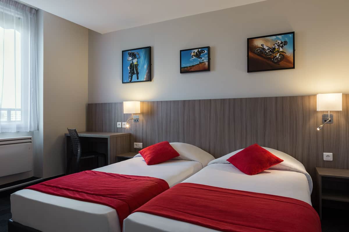 A hotel room with two beds, red bedding, a desk, a chair, and three framed pictures on the wall.