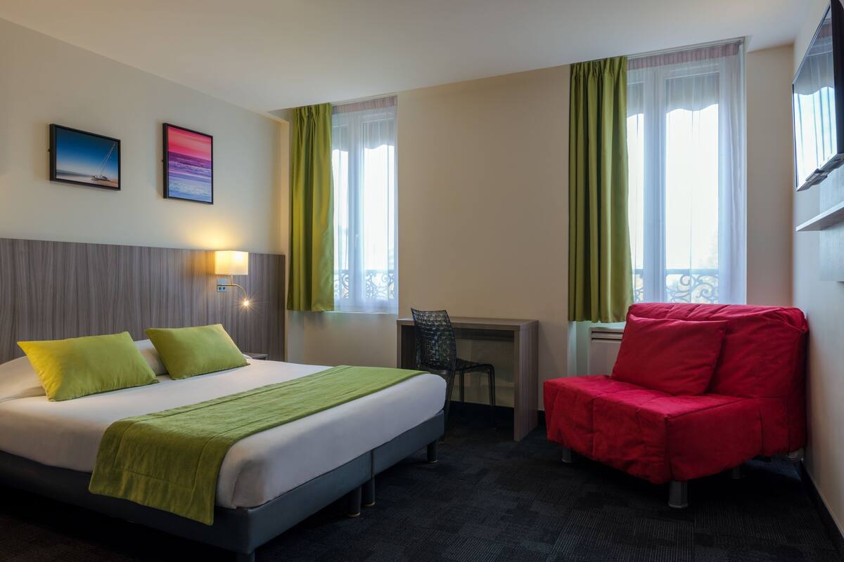 A hotel room with a bed, desk, chair, and two windows with curtains.