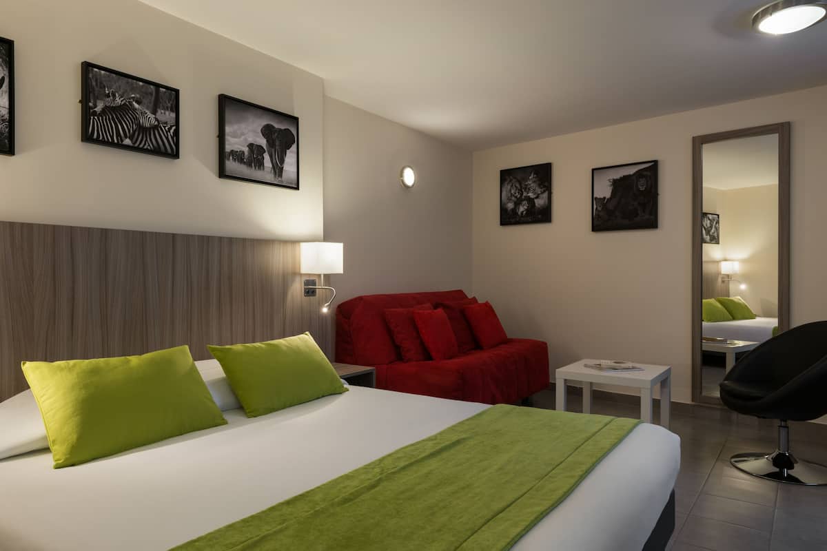 A hotel room with a large bed, a red sofa, a small table, and a chair. There are wall-mounted lamps and framed pictures on the walls.