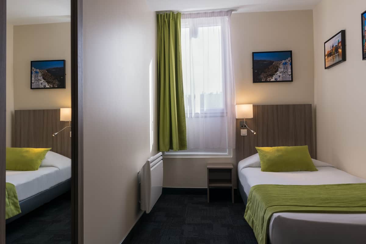 A hotel room with two beds, a window with green curtains, a small table, and wall-mounted lamps.