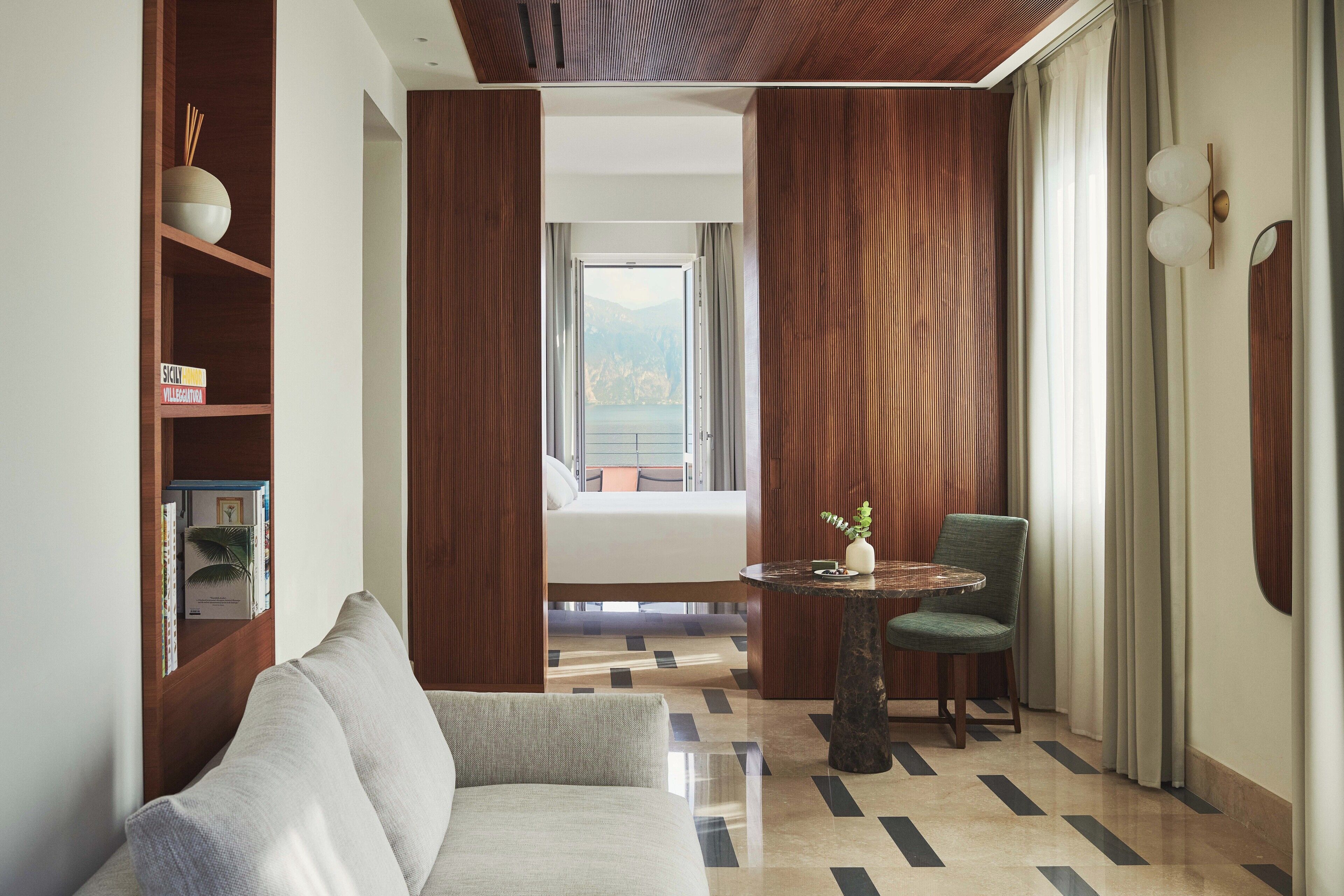 A modern hotel room with a sofa, a small round table with a chair, a bookshelf, and a view of the outdoors through a large window.