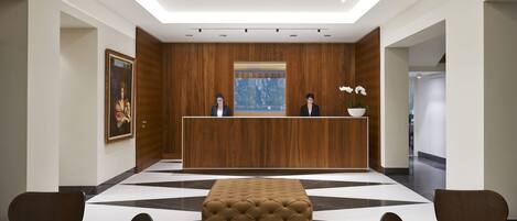A reception area with a wooden reception desk, two seated individuals, a large window, and a checkered floor pattern.