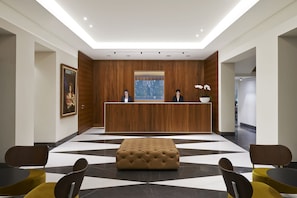 A reception area with a wooden reception desk, two seated individuals, a large window, and a checkered floor pattern.