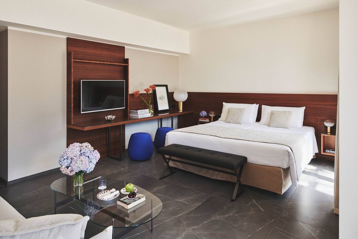 A modern hotel room with a large bed, a flat-screen TV, a glass coffee table, and a bench.
