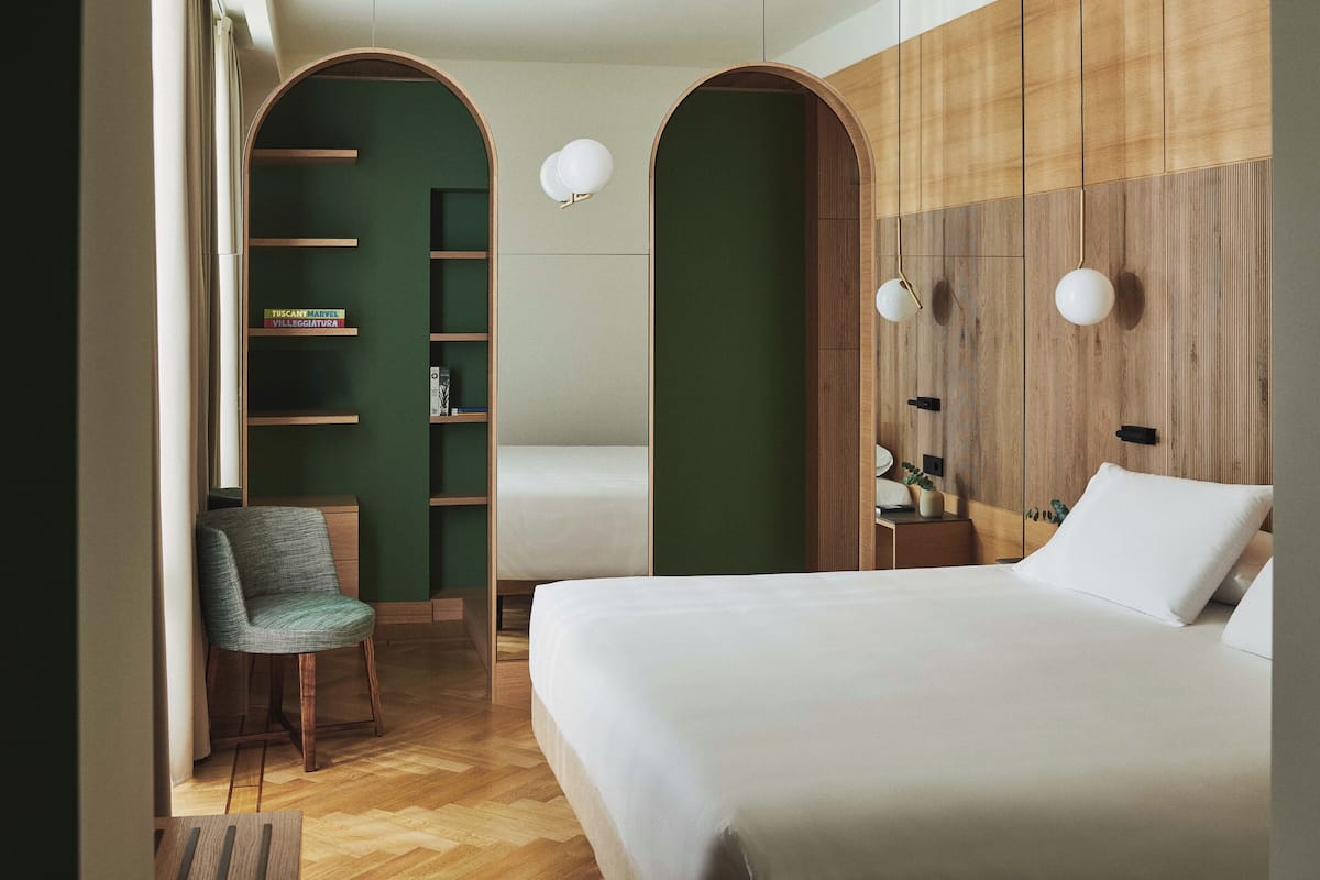 A hotel room with a bed, a chair, a shelf with books, and a wooden wall panel.