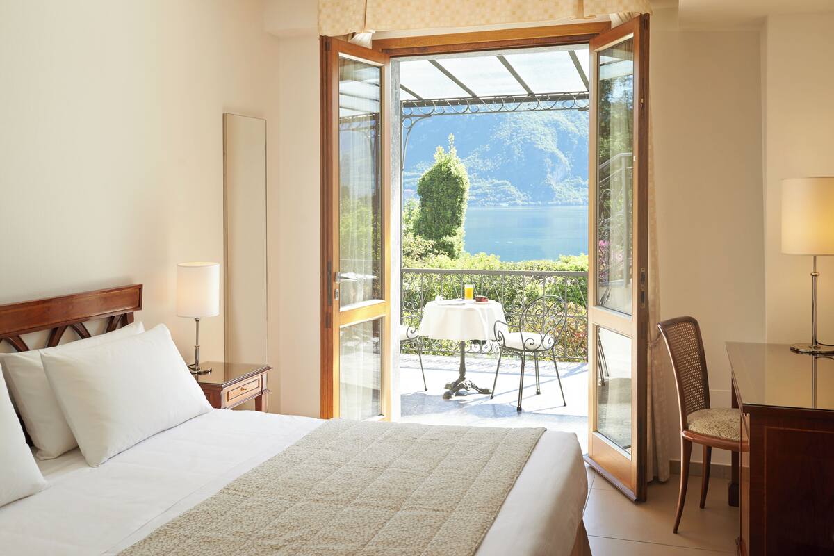 A hotel room with a bed, a nightstand, a table with chairs, and a view of a lake through an open door.