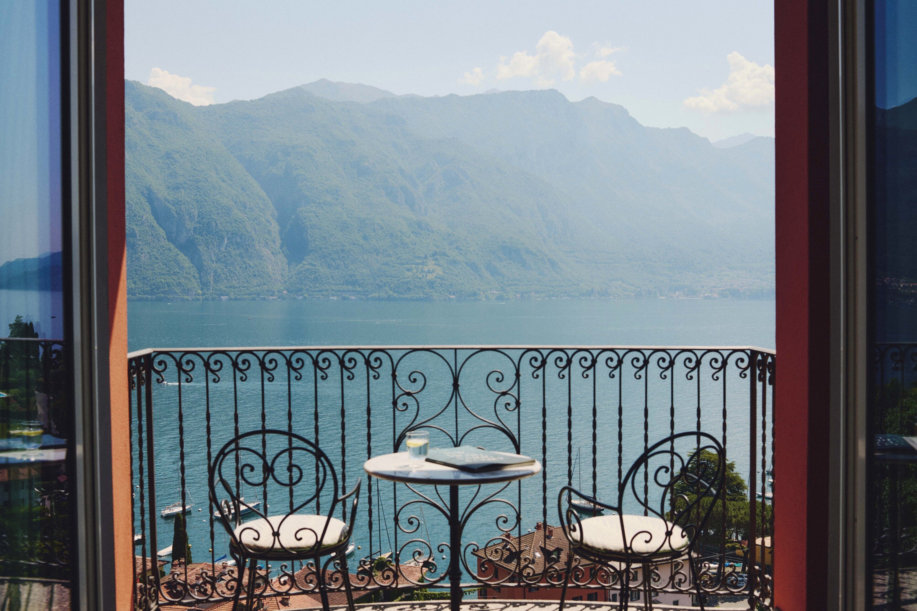 A balcony with a view of a lake and mountains. There are two chairs and a small table.