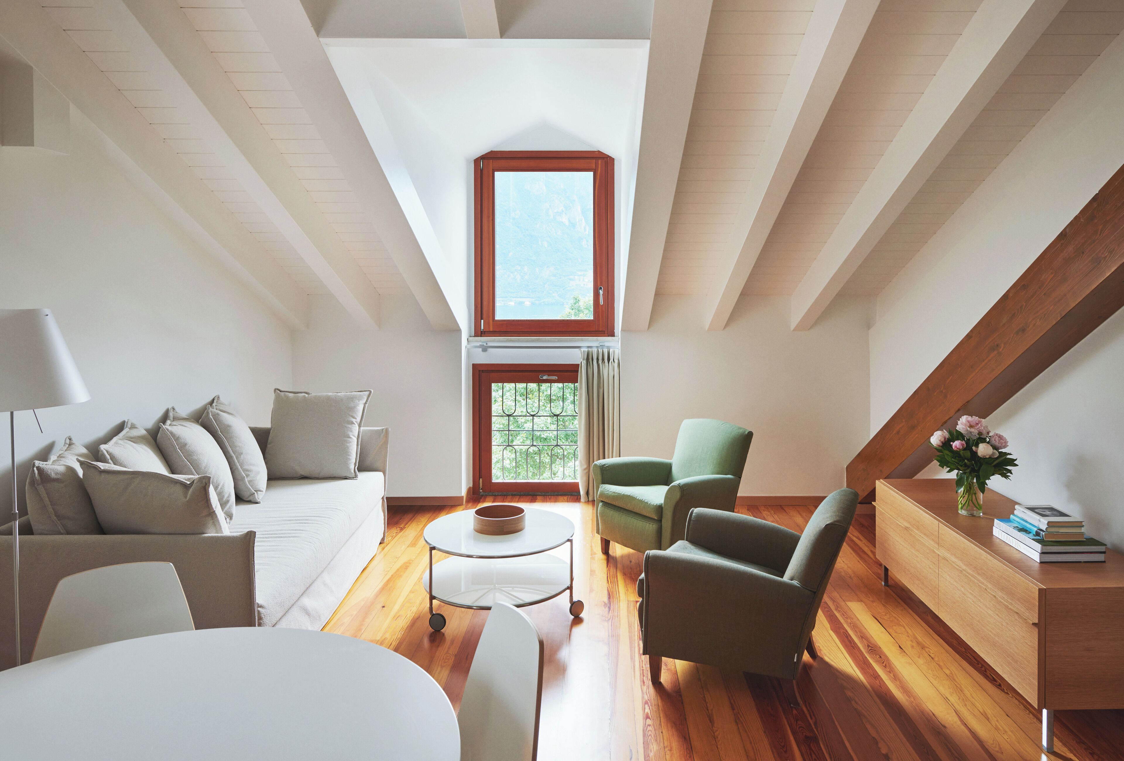 Une chambre de style loft moderne, avec un plafond en pente, une grande lucarne, un plancher en bois, un canapé, deux fauteuils, une petite table ronde et une commode en bois sur laquelle est posé un vase de fleurs.