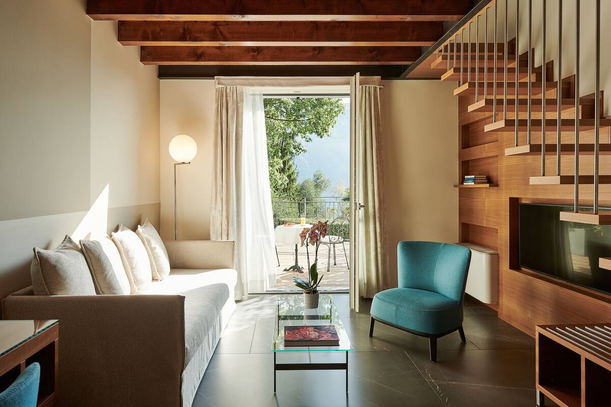 A modern living room with a sofa, a glass coffee table, a teal armchair, a TV, and a wooden staircase.