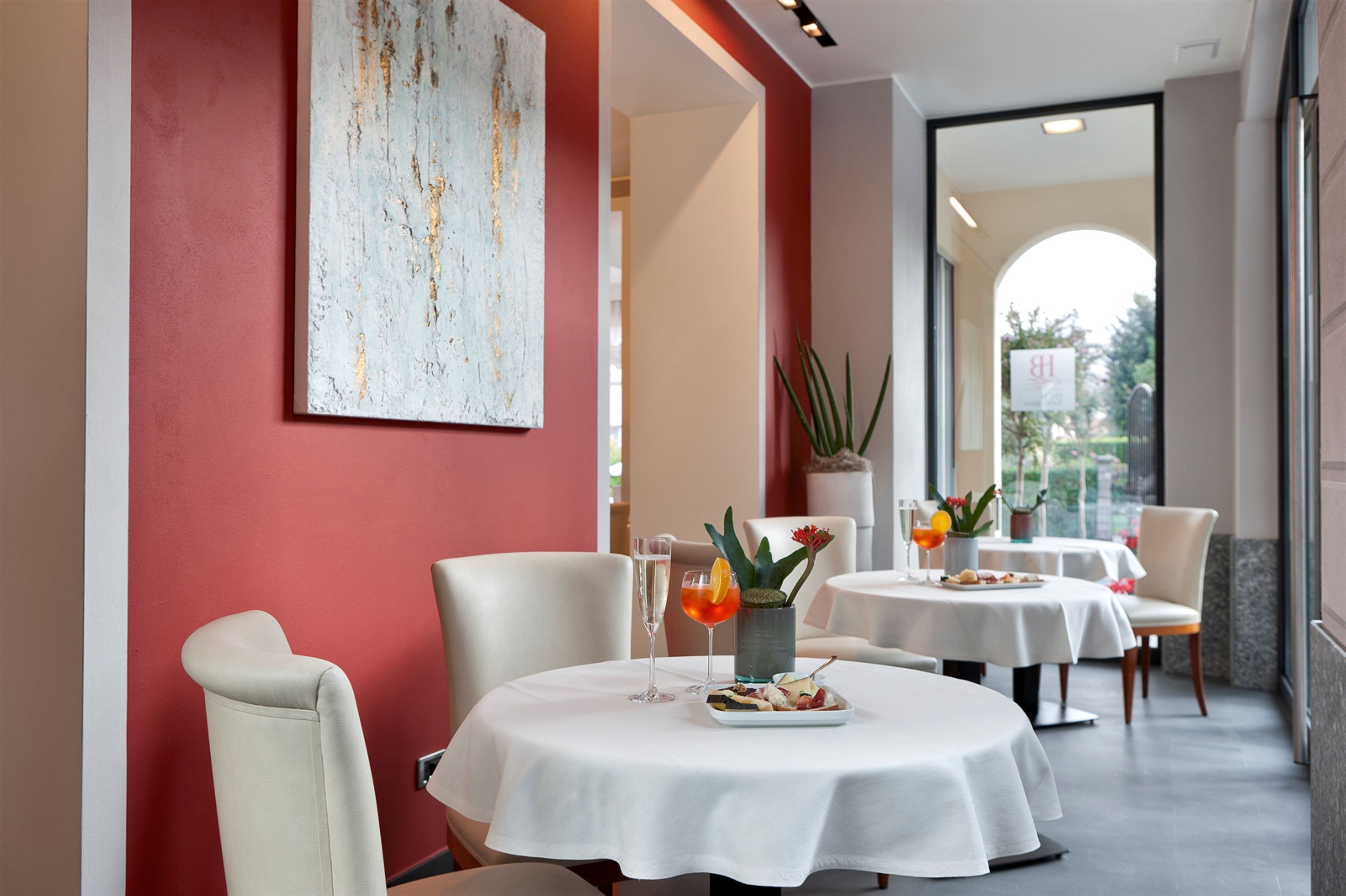A dining area with round tables, white tablecloths, and chairs. There is a large abstract painting on the wall, a potted plant, and a glass of orange juice on one of the tables.