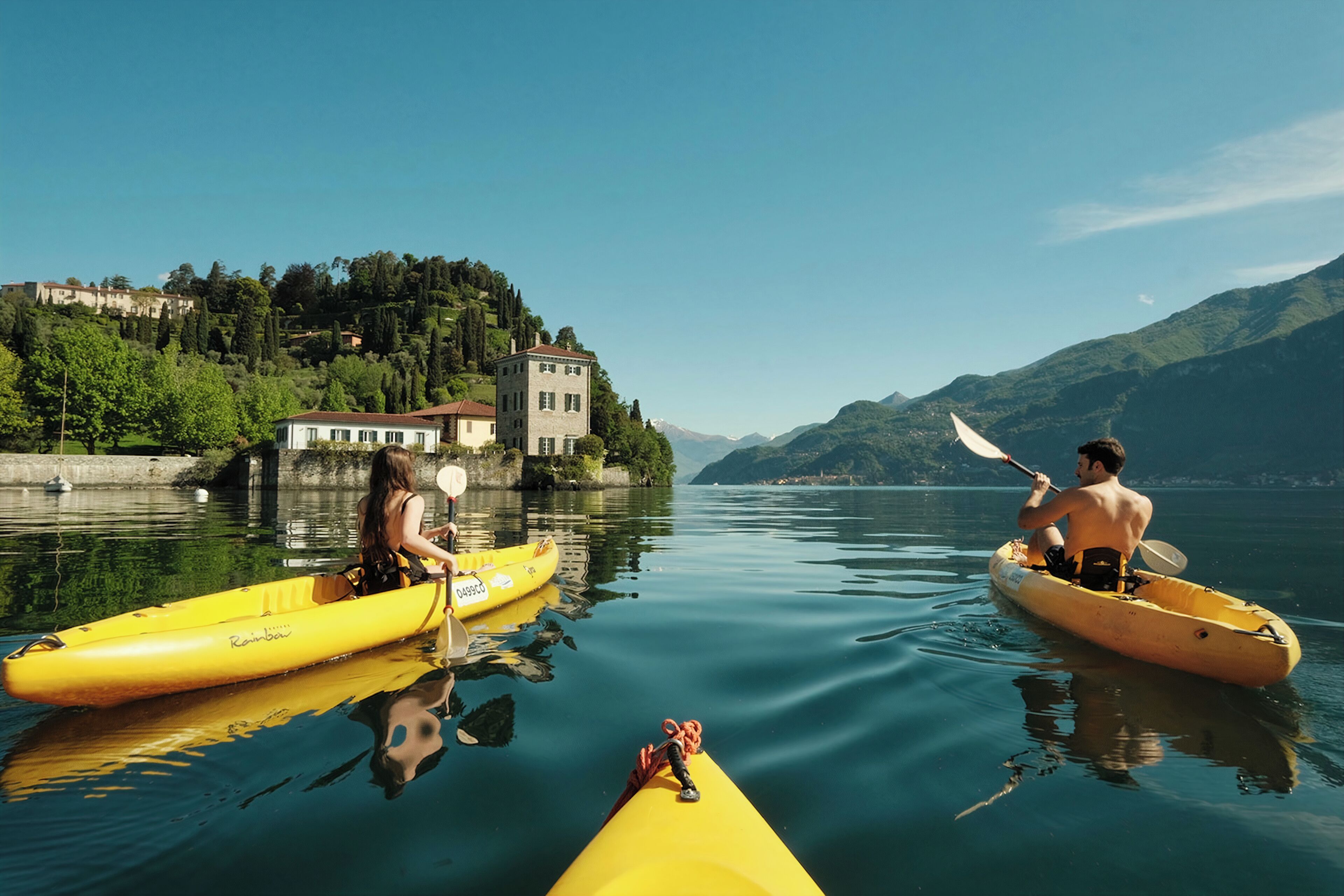 Deux personnes pratiquent le kayak sur un lac paisible, avec une maison et des montagnes en arrière-plan.