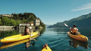 Deux personnes pratiquent le kayak sur un lac paisible, avec une maison et des montagnes en arrière-plan.
