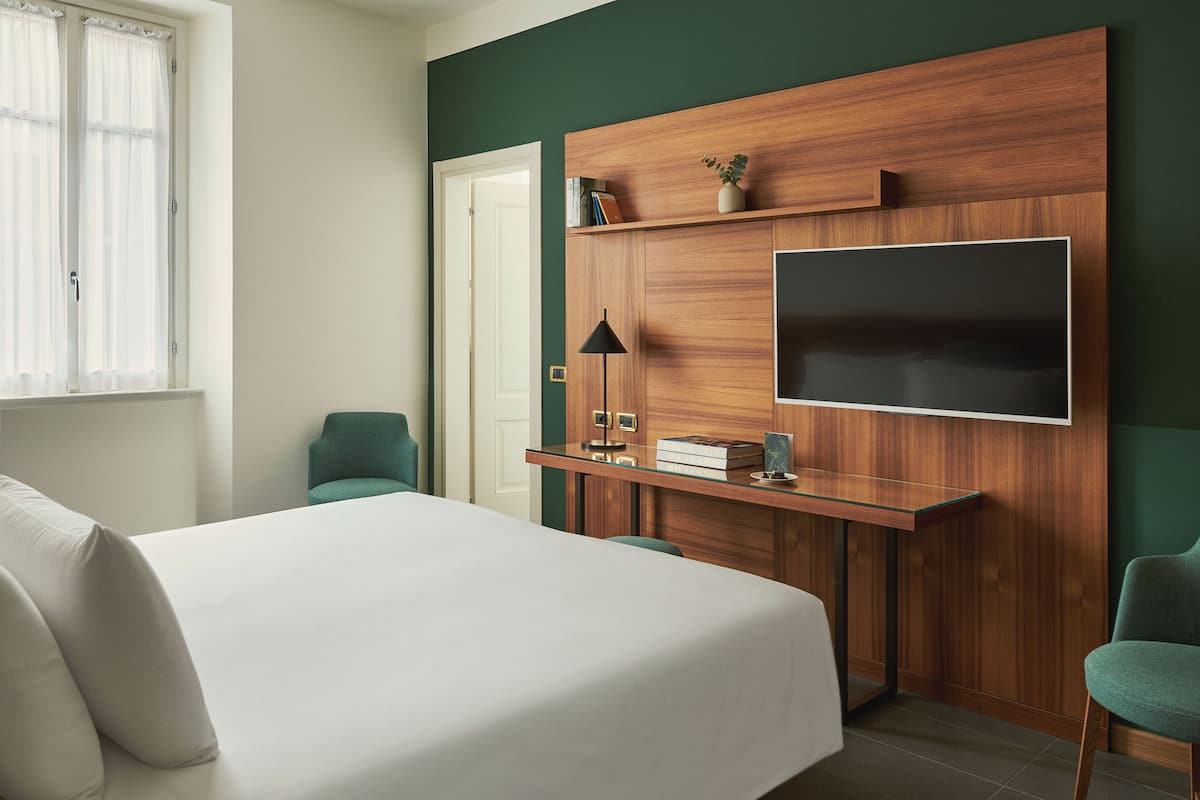 A hotel room with a bed, a desk with a TV, a chair, and a window with curtains.