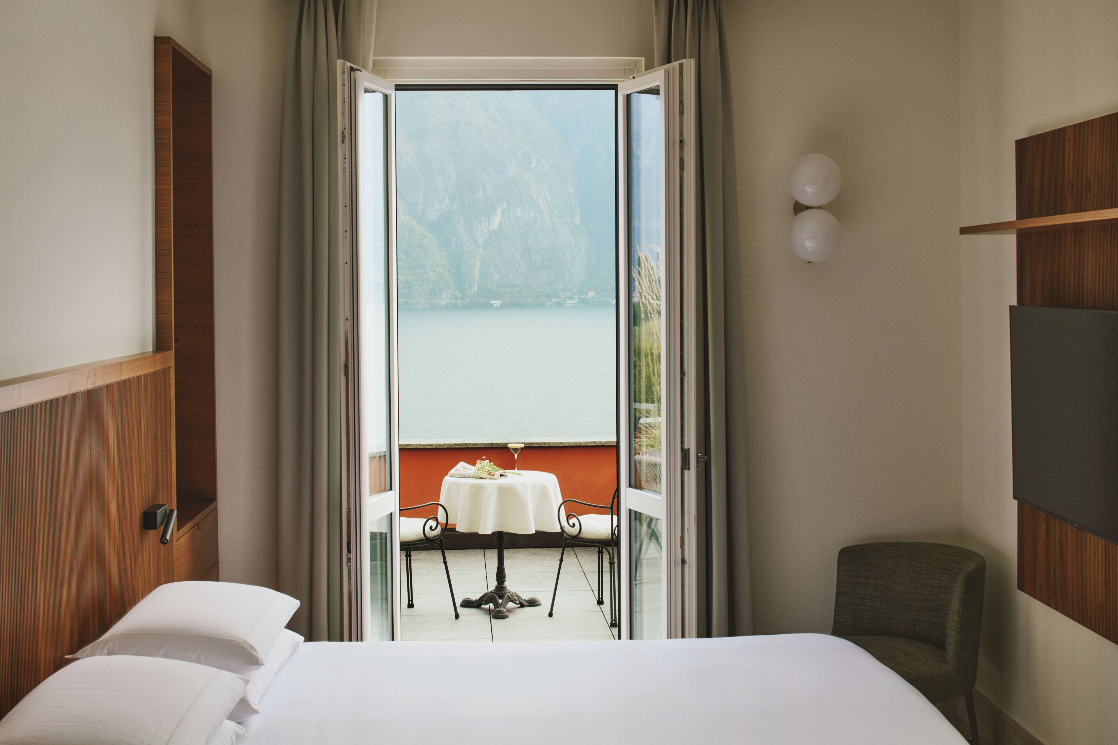 Ein Hotelzimmer mit Bett, einem Tisch mit Stuhl und Blick auf einen See durch eine geöffnete Tür.