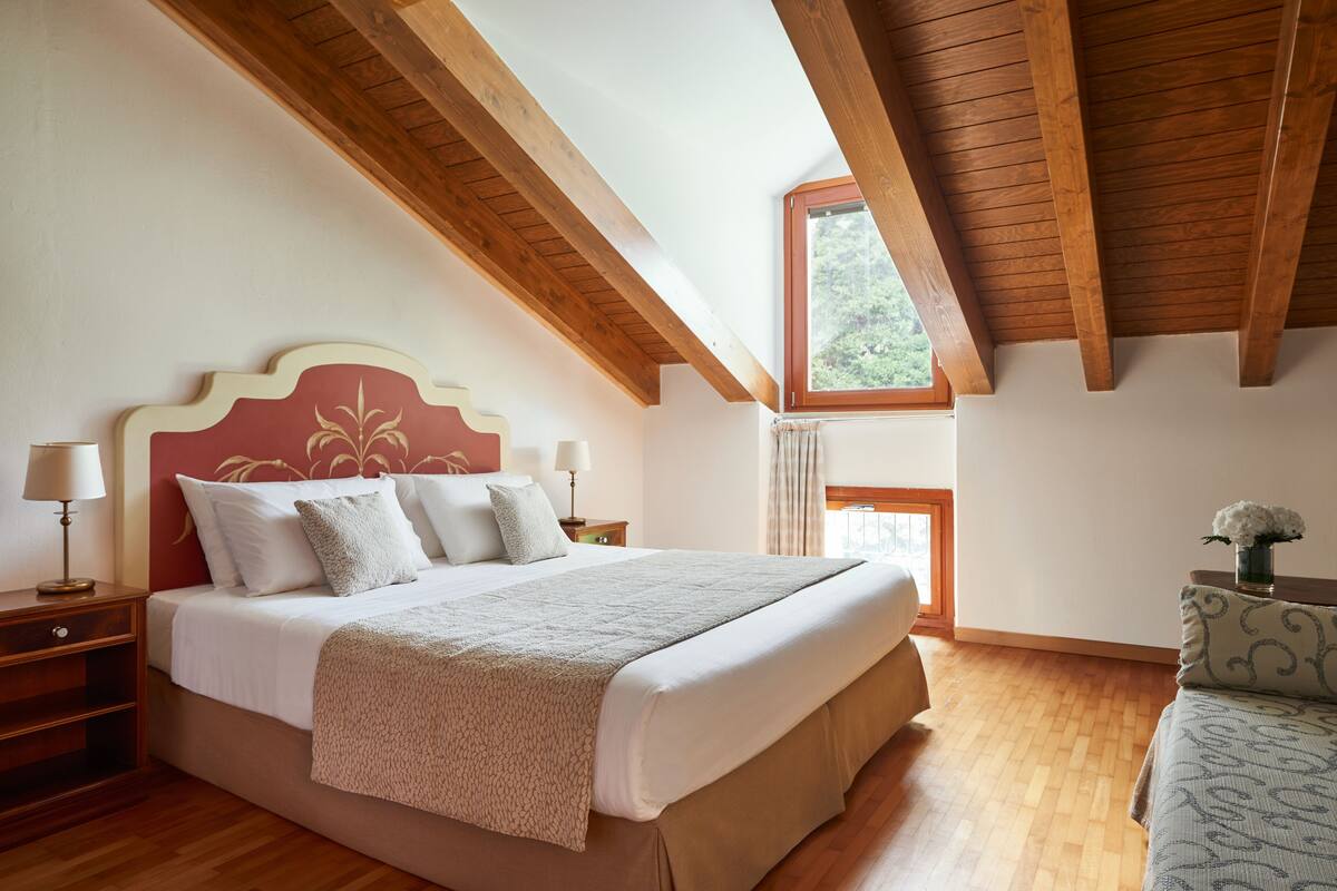 A bedroom with a large bed, wooden beams, a window, and a nightstand with a lamp.