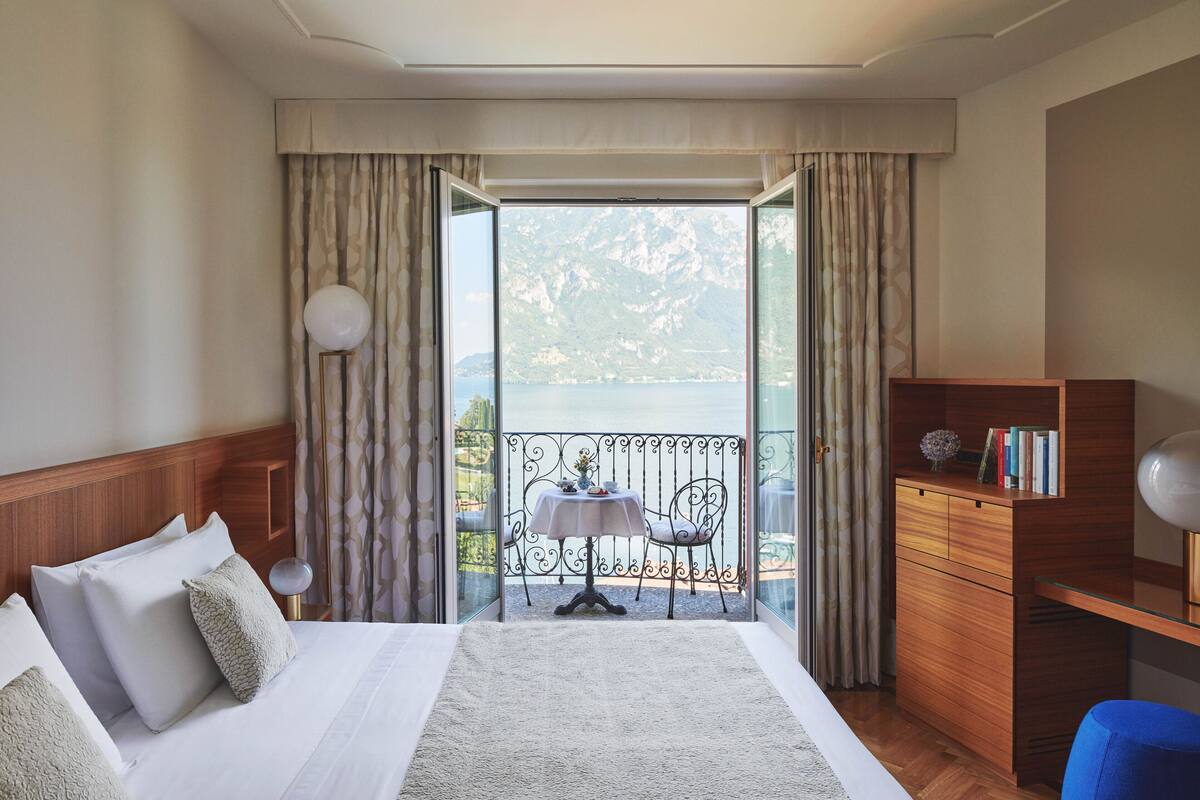 A hotel room with a bed, a desk, a chair, a table, and a view of a mountainous landscape through a balcony.