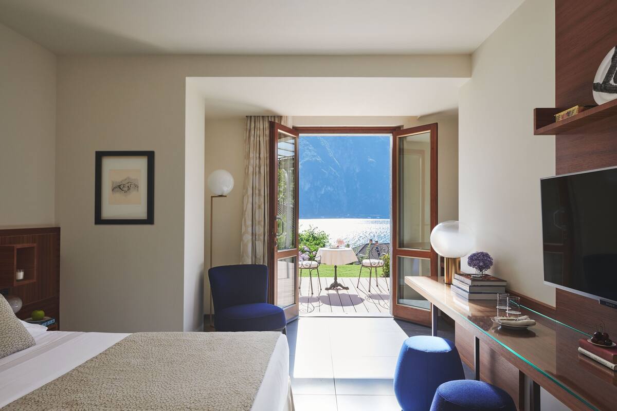A hotel room with a bed, a desk with a TV, a chair, and a view of the sea through an open door.