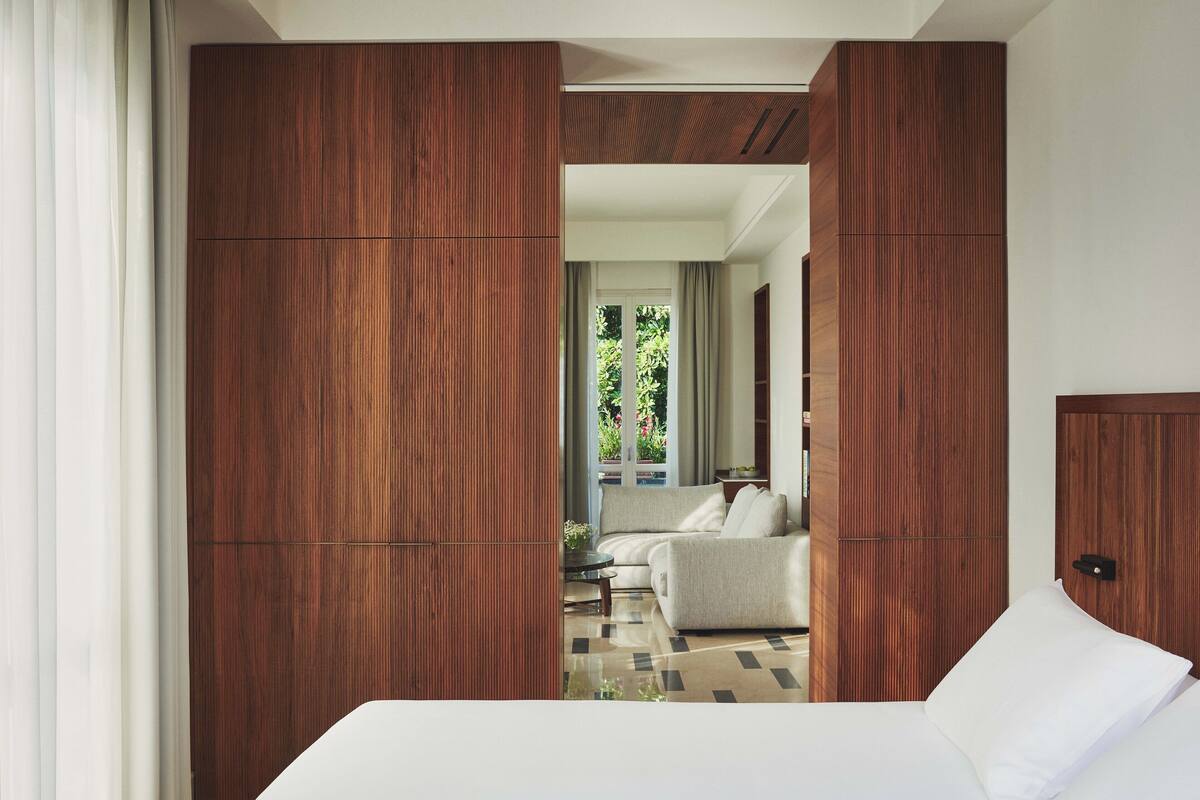A modern hotel room with a large bed, a sofa, and a view of the outdoors through a wooden partition.