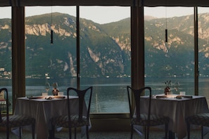 Un comedor con mesas dispuestas para dos personas, con vista a un lago y montañas al fondo.