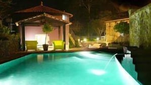 Un área de piscina nocturna con un pabellón, sillas de descanso y una pared de piedra.