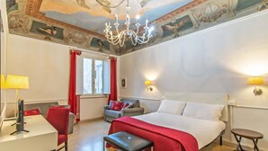 A hotel room with a bed, a desk, a chair, a bench, a chandelier, and a ceiling mural. - Duomo View B&B (Florence)