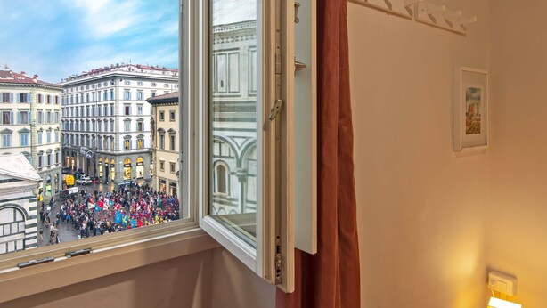A room with a window overlooking a busy street, a bed, a bedside table with a lamp, and a framed picture on the wall. - Duomo View B&B (Florence)