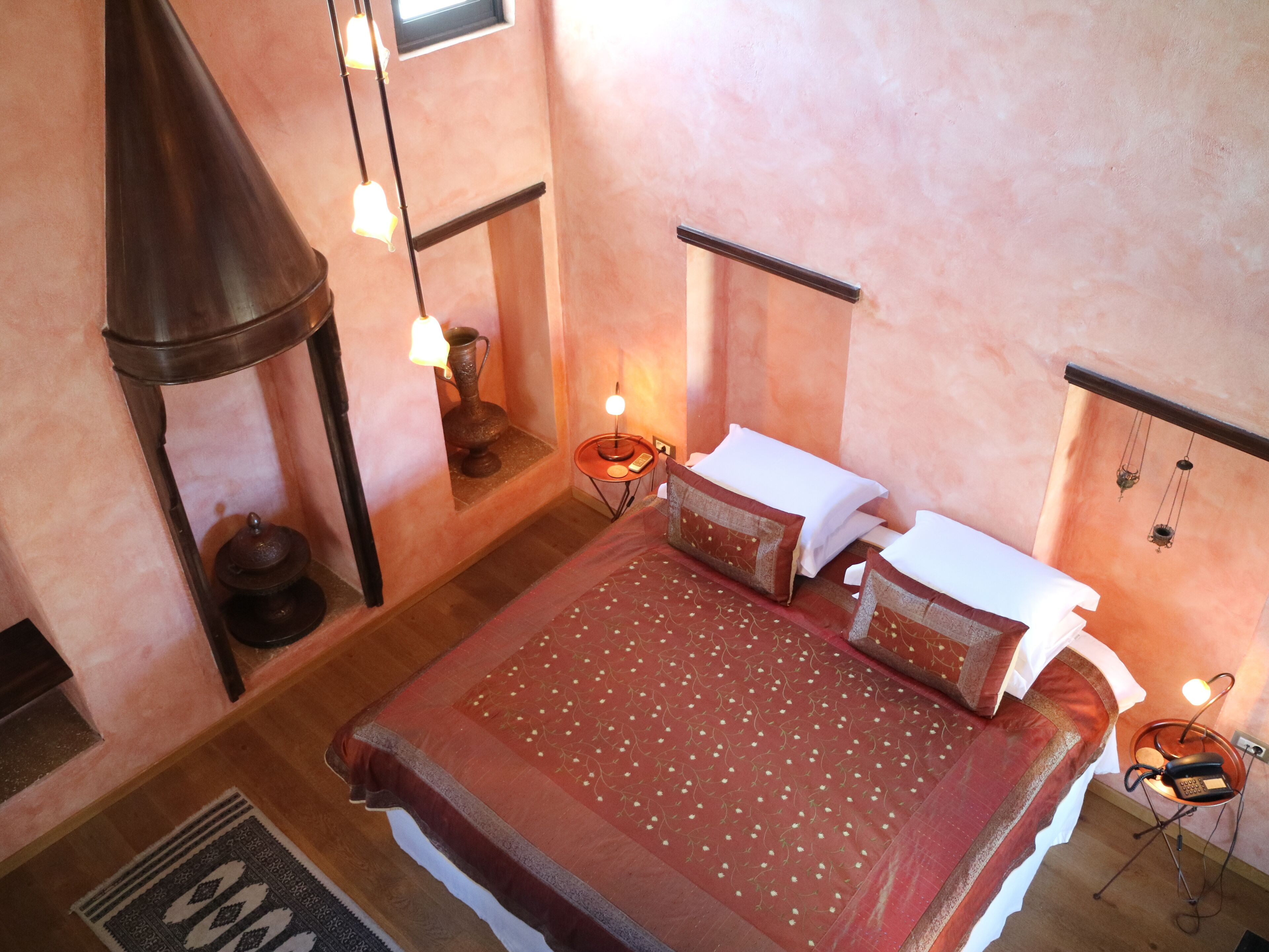A bedroom with a large bed, a hanging light fixture, a small table with a lamp, and a wall-mounted telephone.