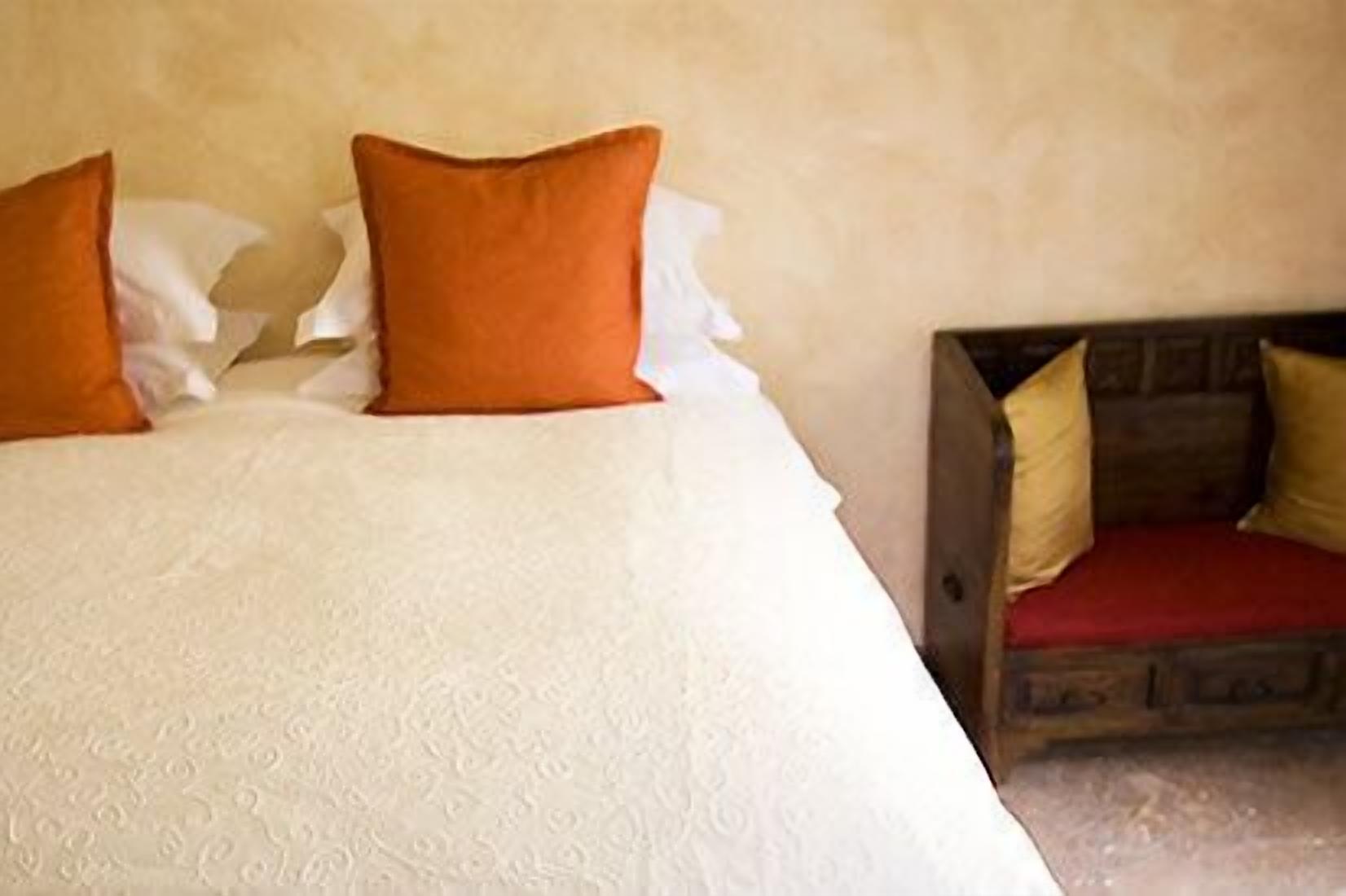 Una cama con ropa de cama blanca, almohadas naranjas, un cabecero de madera y un cojín rojo sobre un banco de madera.