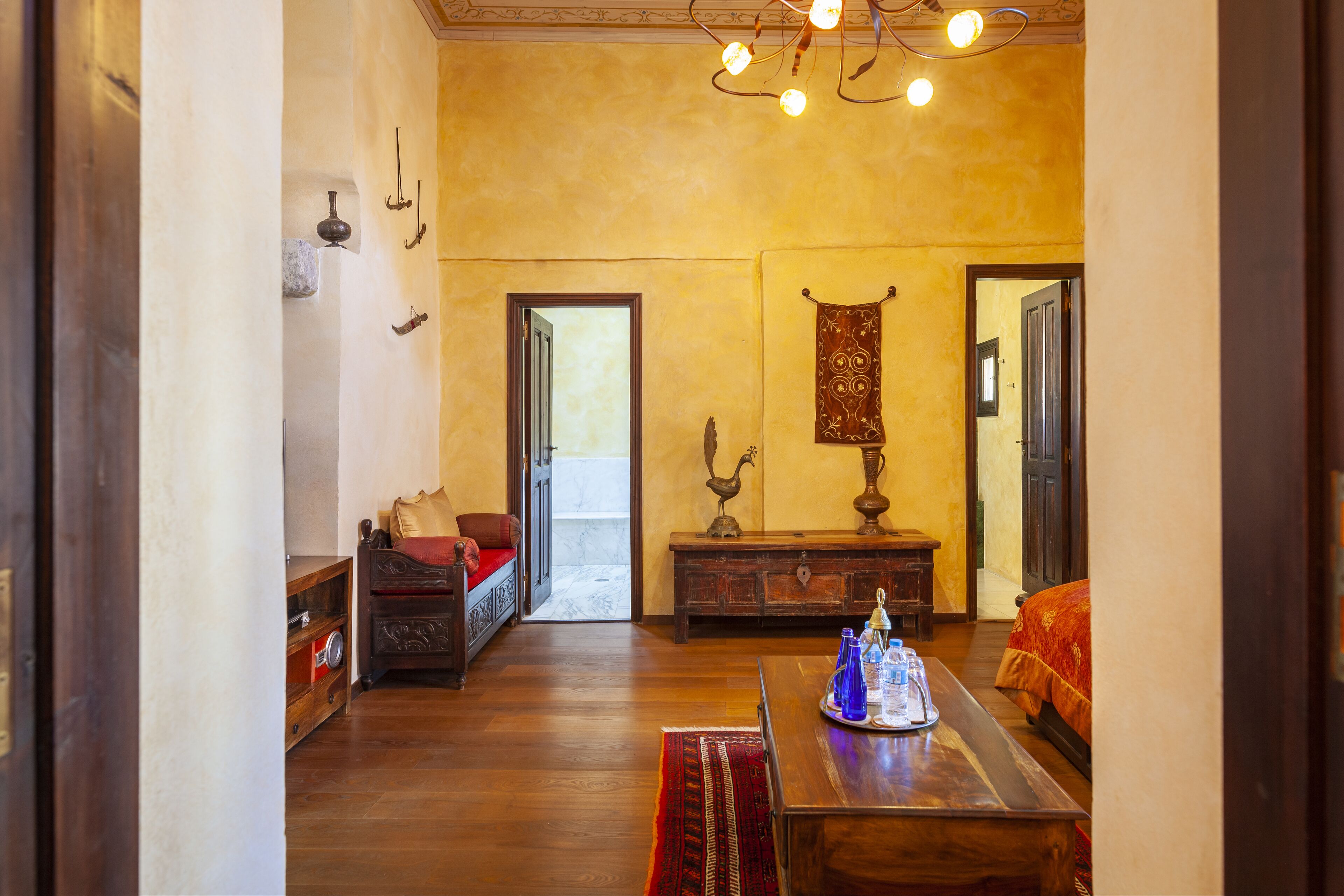 Una habitación con piso de madera, una cama con manta roja, una mesa de madera con una bandeja y un tapiz colgante.