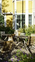 Un espacio al aire libre con mesas y sillas de madera, rodeado de follaje y un edificio con amplias ventanas.