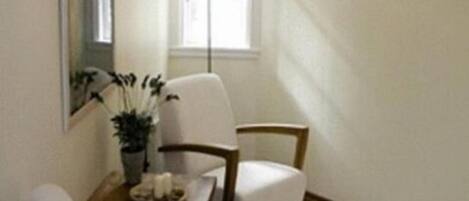 A room with a white armchair, a wooden side table with a plant, and a small table with a lamp.