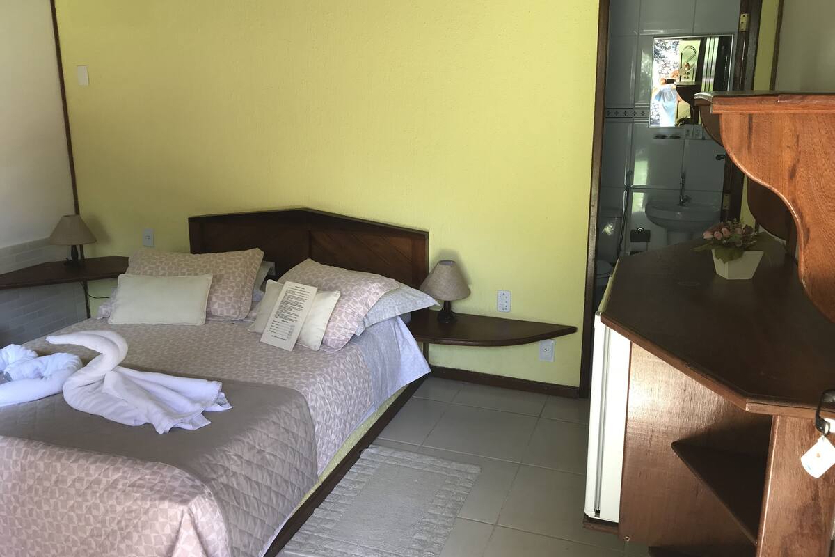A hotel room with a bed, bedside table, lamp, and a small wooden cabinet.