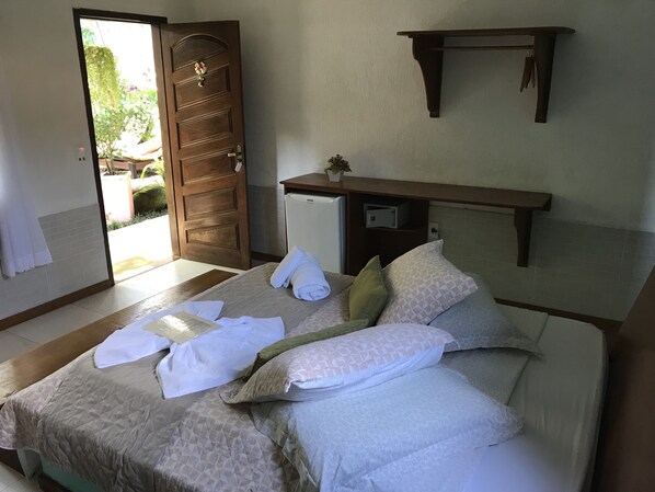 A neatly made bed with pillows and towels, a small refrigerator, and a wooden shelf above it.