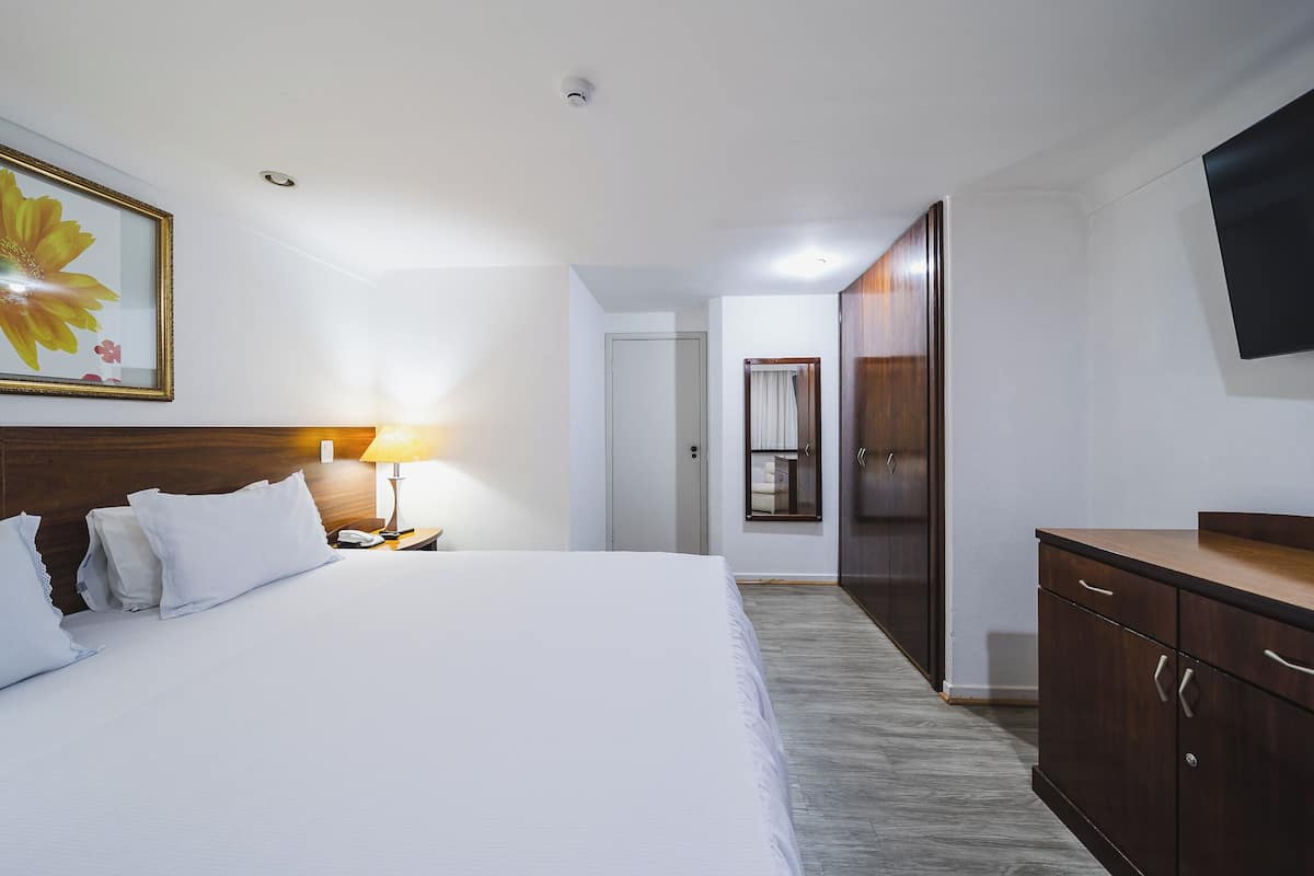 Habitación de hotel con cama, mesita de noche, lámpara, armario y televisor montado en la pared.