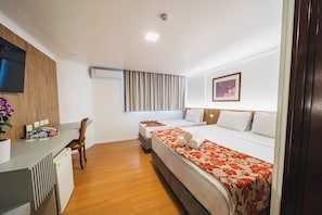 A hotel room with a desk, two beds, a TV, an air conditioner, and a window with curtains.