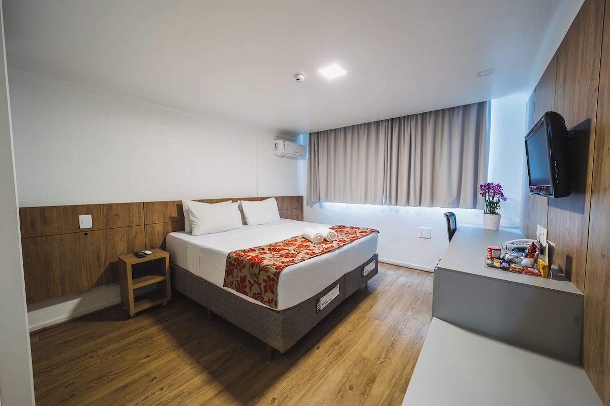 Una habitación de hotel moderna con cama, mesita de noche, televisión y ventana con cortinas.