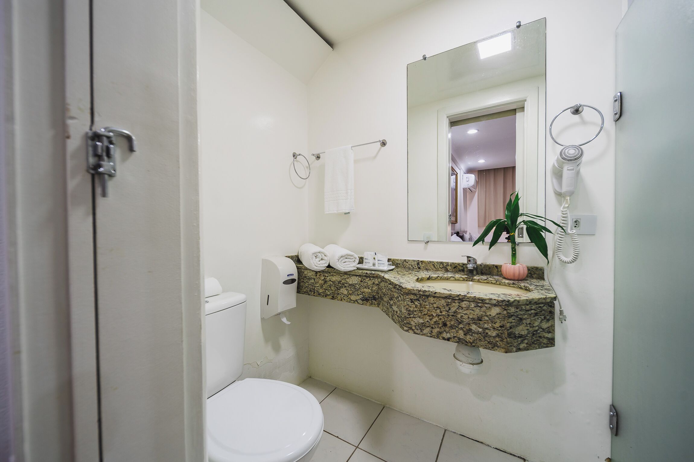 Casa de banho com sanita, lavatório, espelho, dispensador de toalhas e secador de cabelo.