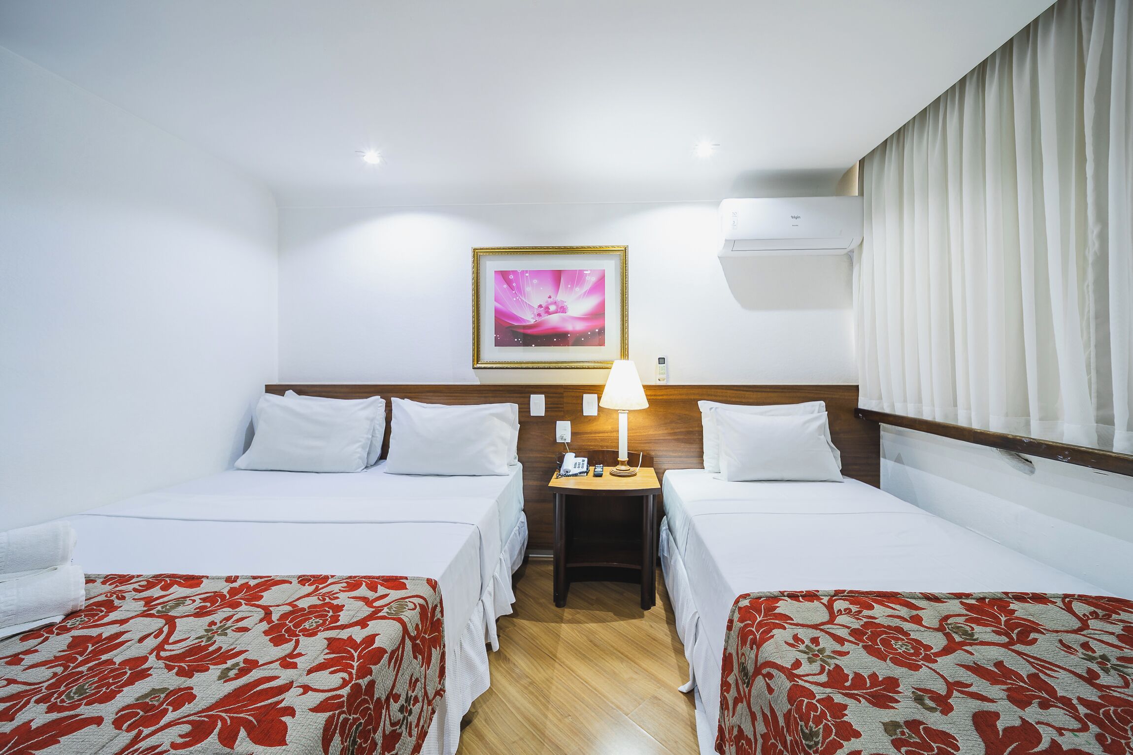 A hotel room with two beds, a nightstand with a lamp, an air conditioning unit, and a framed picture on the wall.