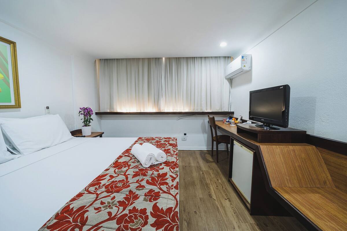 Una habitación de hotel con cama, escritorio, computadora, televisor y ventana con cortinas.